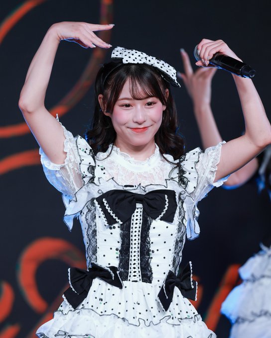 A young woman performing on stage in a black and white polka-dot dress with ruffled sleeves and bow accents, making a heart shape with her arms while holding a microphone.