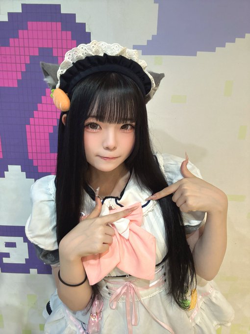 A young woman in a white and pink maid outfit with cat ears poses with finger hearts against a pixelated background.
