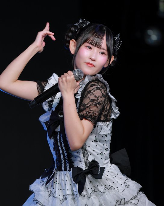 A young performer on stage holding a microphone, wearing a black and white gothic-lolita style dress with lace details, polka dots, and black bows, along with black cat-ear hair accessories.