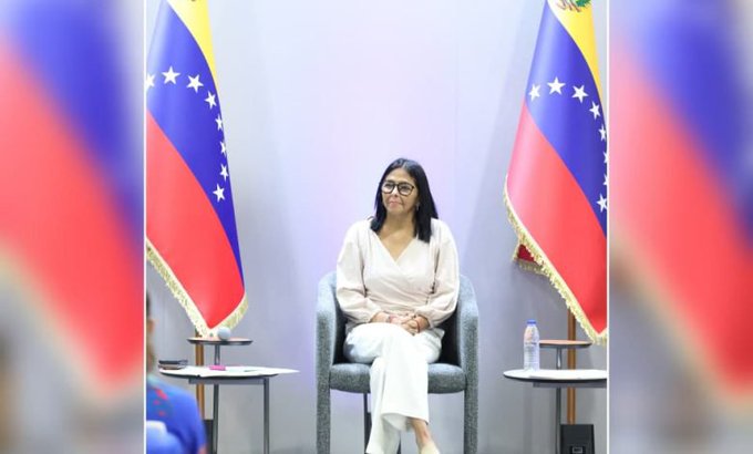 A woman with dark hair and glasses wearing a white outfit sits in a gray chair between two Venezuelan flags.