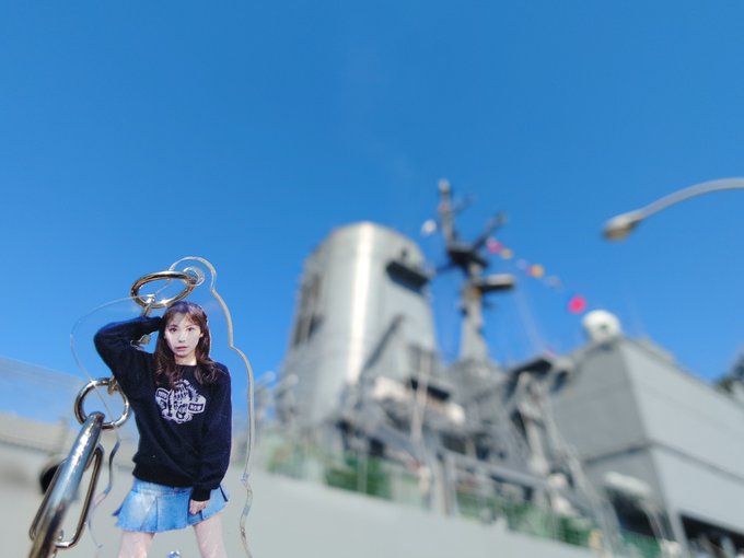 A young woman in a black hoodie and denim skirt poses outdoors with a clear balloon, with a large naval ship visible in the background.
