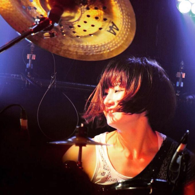 A drummer with short dark hair and reddish highlights performs under stage lights, smiling beneath a large golden cymbal.