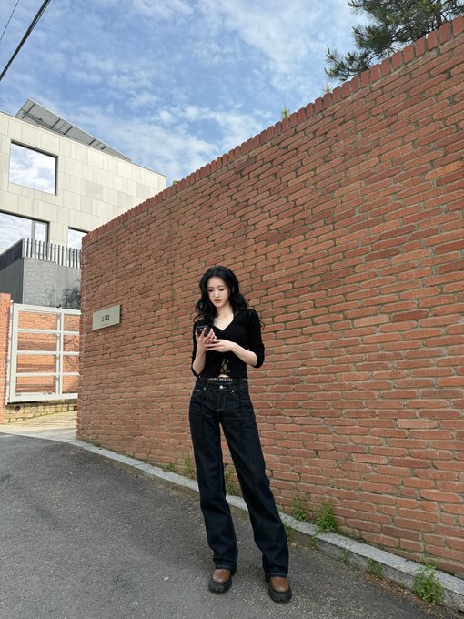 A young woman with long black hair stands against a red brick wall, dressed in an all-black outfit of a cropped cardigan and dark jeans, looking down at her phone while the date "260421" (April 26, 2021) likely commemorates when the photo was taken. The "212" house number plaque is visible on the wall beside her, adding a subtle detail to the urban scene.