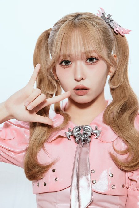 A young woman with blonde pigtails wearing a pink vinyl outfit with a heart-shaped brooch, posing with a hand gesture near her face.