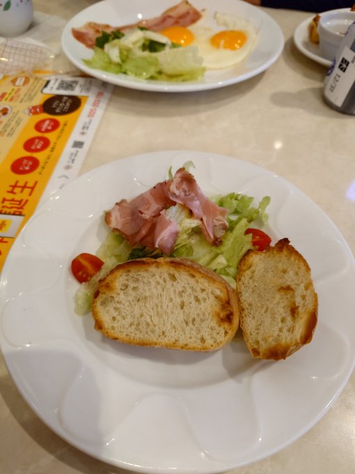 A plate with two slices of toasted bread, ham, and a small salad, with a Japanese breakfast set including fried eggs and bacon in the background.