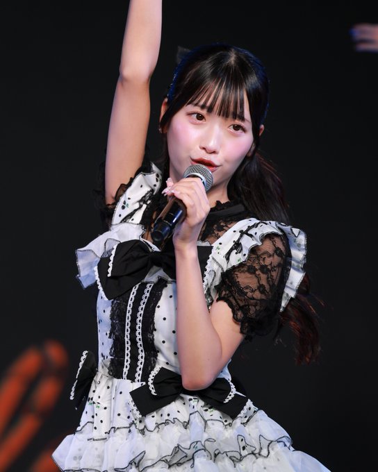 A young Japanese performer singing into a microphone while wearing a black and white lace dress with bows, performing on a dark stage.