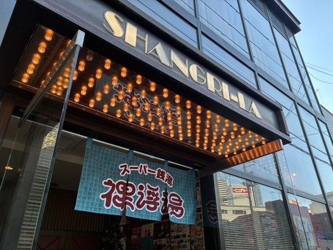 The image shows the exterior entrance of Shanchiglia (シャングリラ) in Osaka's Umeda district, decorated with illuminated marquee bulbs and a hanging banner that reads スーパー銭湯「裸浮湯」(Super Sento "Hadakabuyu"). The venue's striking facade sets the stage for the third Super Sento "Hadakabuyu" event, with merchandise sales opening at 11:00 before the show.