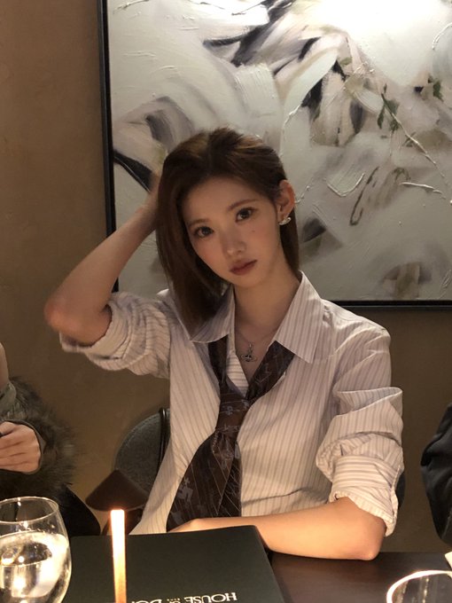A young woman in a striped white shirt and brown patterned tie poses with her hand in her hair at a dimly lit restaurant, with an abstract painting behind her and a lit candle on the table.