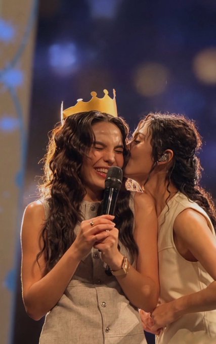 Two women are on stage; one wearing a paper crown holds a microphone with her eyes closed, while the other leans in to kiss her cheek.