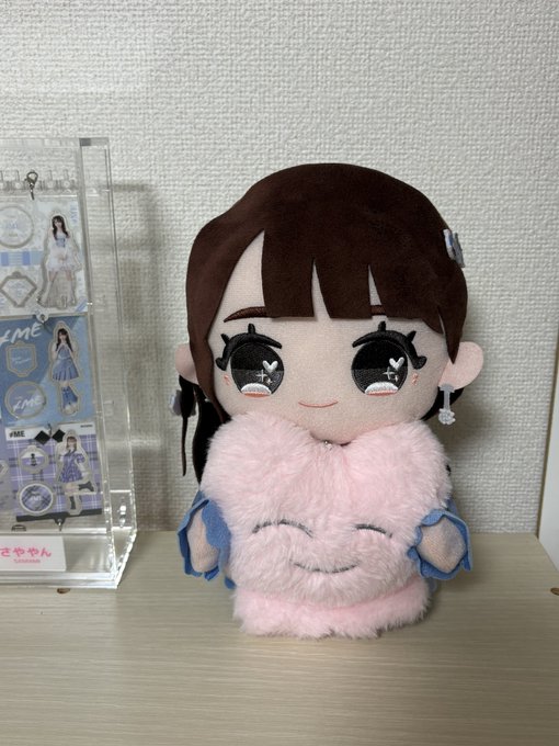 A plush doll of a girl with brown hair holds a smaller pink fluffy bunny plush, sitting next to an acrylic display stand featuring character merchandise.