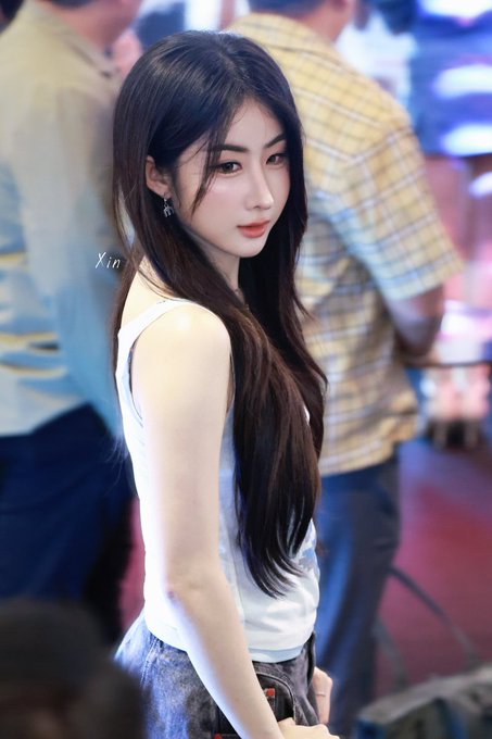 A photograph of a young woman with long dark hair wearing a white tank top, taken at what appears to be an event or convention.