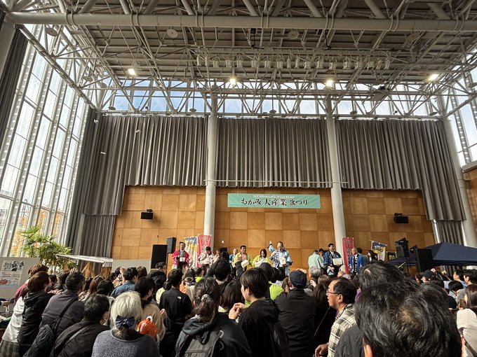 A photo of a crowded indoor event at a large venue with high ceilings and tall windows, where a group of people in traditional Japanese attire are performing on stage under a banner reading "おおがき産業まつり" (Ogaki Industry Festival).