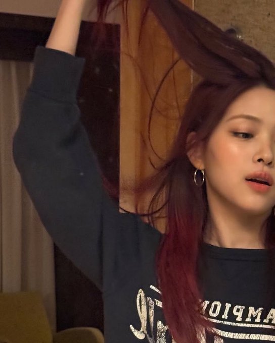 A young woman with long dark hair tinted red raises her arm, wearing a dark sweatshirt with white lettering.