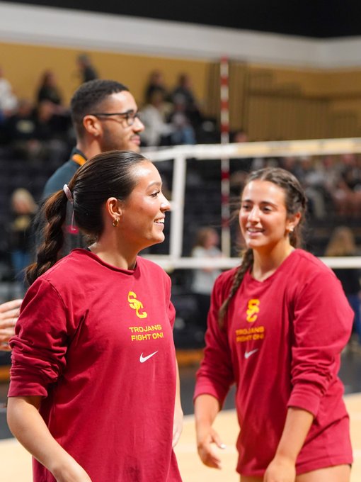 Two smiling female volleyball players wearing red 'Trojans Fight On' USC sweatshirts with Nike logos, standing on a court with a volleyball net and spectators visible in the background.