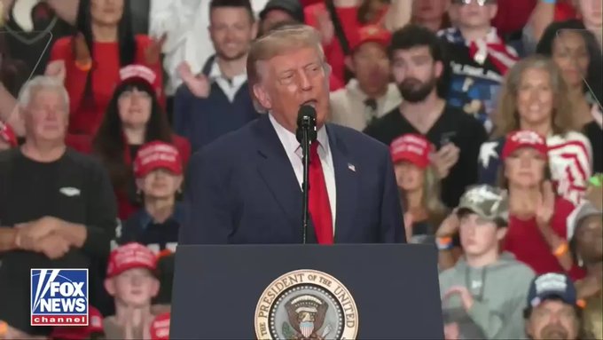 Donald Trump stands at a presidential podium speaking at a rally, with many supporters wearing red 'Make America Great Again' hats in the background and a Fox News Channel logo in the bottom left corner.