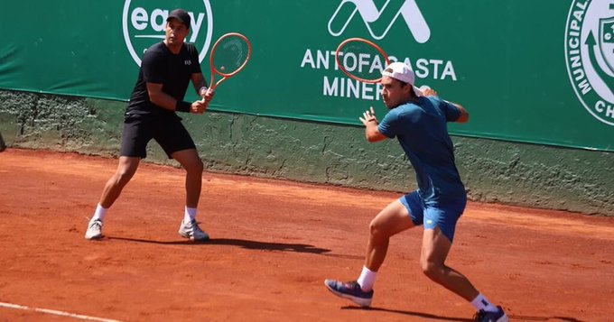 Two male tennis players in action on a red clay court, with sponsor banners for 'ANTOFAGASTA MINERALS' and 'egg' visible on the green backdrop.