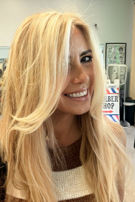 A smiling woman with long blonde hair wearing a brown and cream-colored sweater, photographed indoors with a barber shop pole visible in the background.