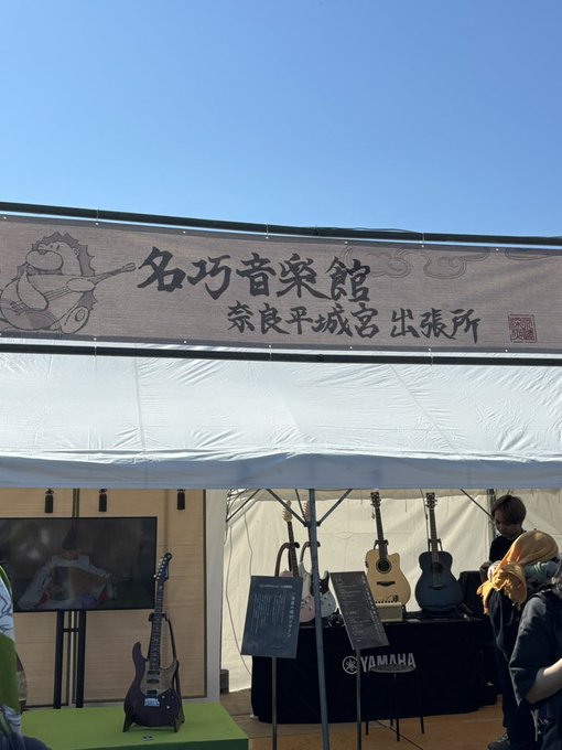 An outdoor tent display for "名巧音楽館" (Nakou Music Hall) at the 奈良平城宮 出張所 (Nara Heijō Palace branch), showcasing Yamaha acoustic and electric guitars and a bass guitar on display.