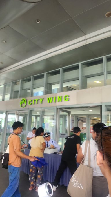 People gather at a 'CITY WING' counter inside a building, including a man in a police uniform and a woman holding a roll of paper.