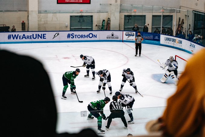 An indoor ice hockey game in progress with players in green and white uniforms, a referee, and a goalie, with banners reading '#WeAreUNE', '#StormOn', 'Langford & Low Construction', and 'PortlandGlass.com' visible.