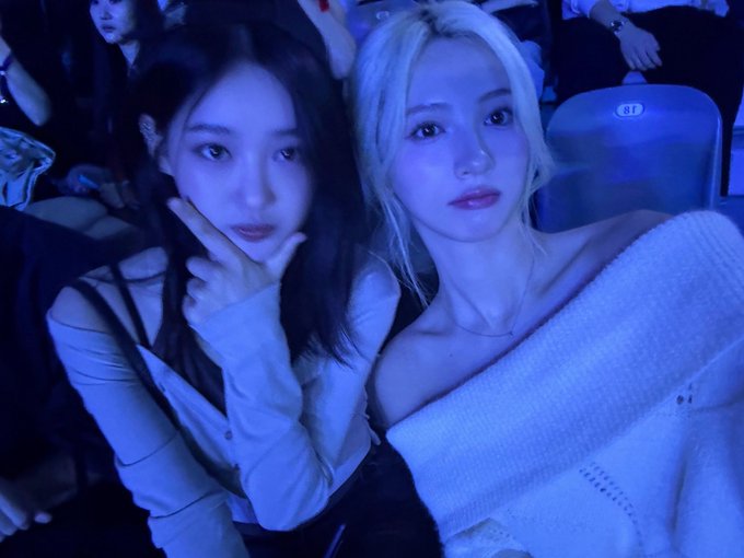 Two young women posing together in a dimly lit venue with blue lighting, one with dark hair and the other with blonde hair.