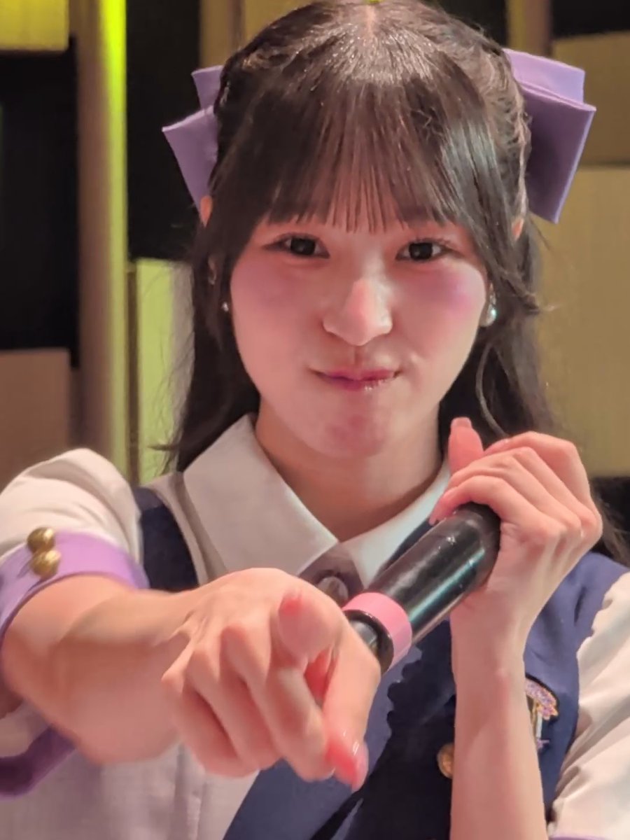 A close-up selfie of a young woman with long dark hair wearing a purple hair accessory and pearl earrings, holding a makeup product toward the camera.