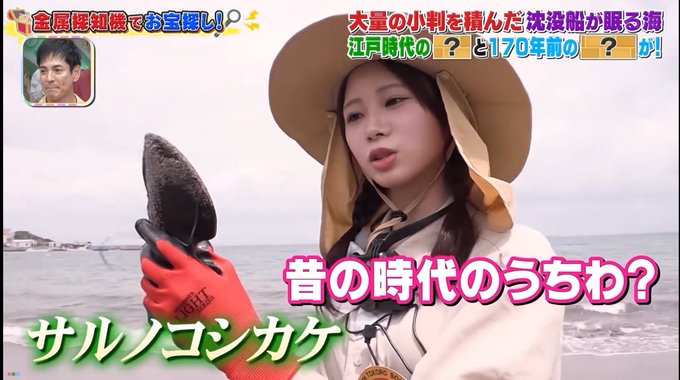 A TV show screenshot featuring a woman in a tan sun hat holding a dark object on a beach, with Japanese text overlaid in pink and green.
