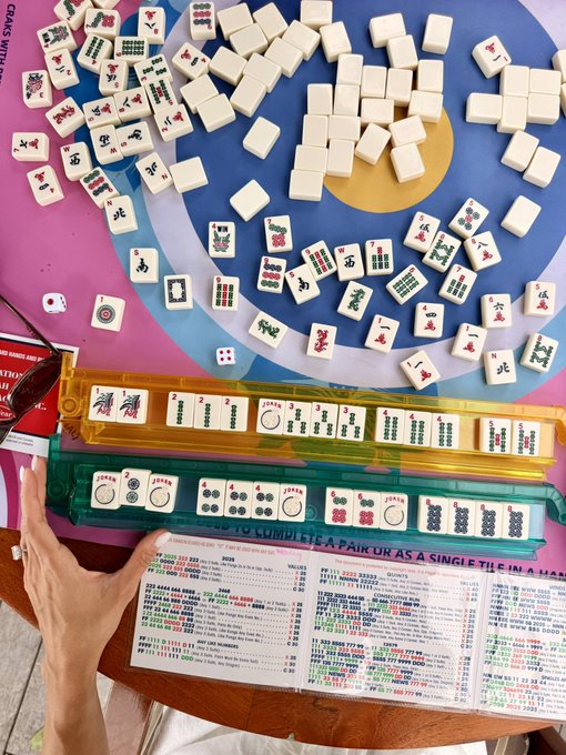 An overhead view of a mahjong game in progress, featuring white and green tiles with Chinese characters and bamboo designs spread across a colorful mat, alongside a printed reference sheet and scorecards.
