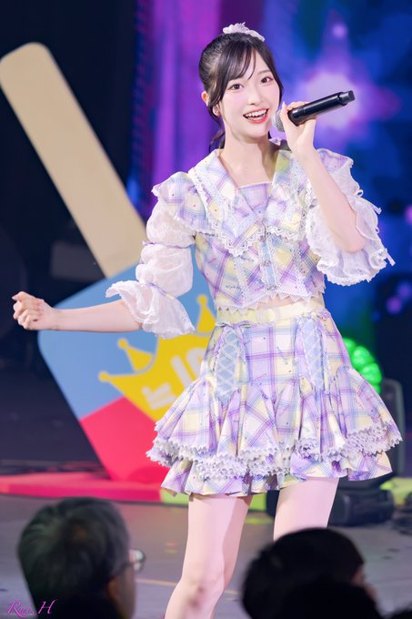 A young female performer in a pastel plaid outfit smiles brightly while singing into a microphone on stage.