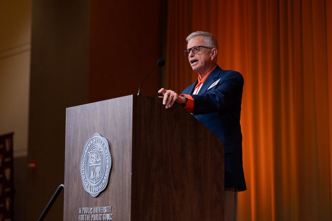 An older man with glasses speaks at a wooden podium bearing the Georgetown University seal, set against an orange curtain backdrop.