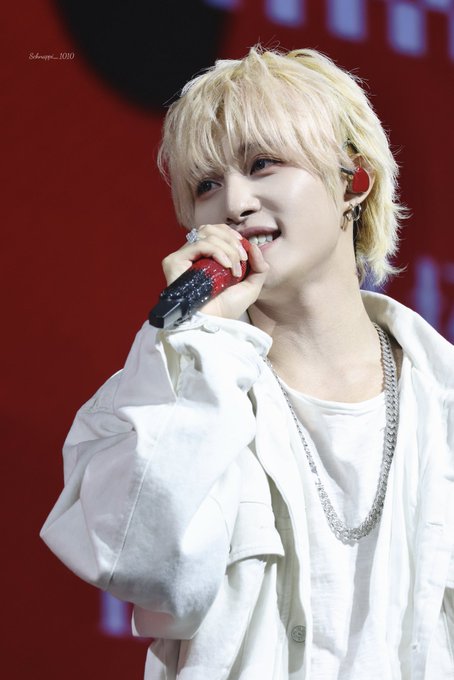 A blonde-haired K-pop performer with an in-ear monitor sings into a red microphone while wearing a white outfit and silver chain necklace, performing on stage.