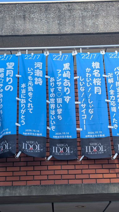 A row of blue vertical banners hanging on a brick wall, each marked '22/7' and 'SHIGA IDOL COLLECTION 2026.10.10' with handwritten Japanese messages.