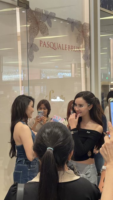 A group of young women pose for a photo in front of a glass storefront for the brand "PASQUALE BRUNI," with one woman in a black off-shoulder top applying lipstick while others watch.