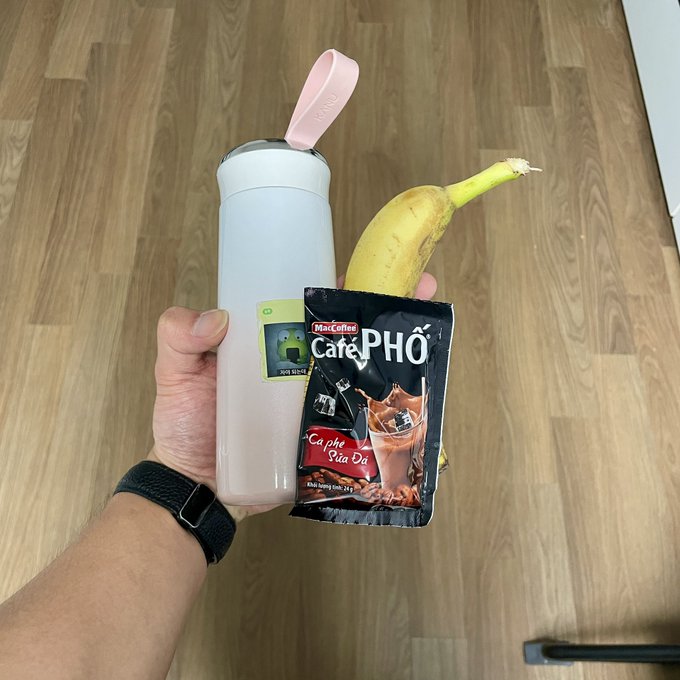 A hand holds a pink Owala water bottle, a banana, and a packet of Mǎ Coffee Café Phố Vietnamese iced coffee with milk, photographed against a wooden floor background.