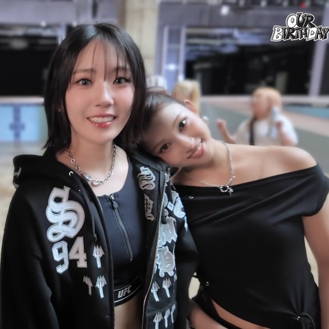Two young women pose closely together for a photo in a large indoor space, with the text 'OUR BIRTHDAY' overlaid in the upper right corner; one wears a black hoodie with '94' and 'UFC' graphics.