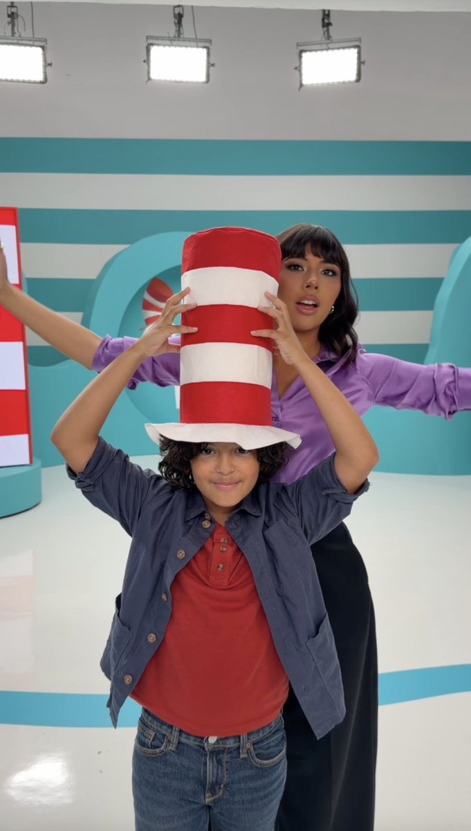 A young boy balances a tall red-and-white striped Cat in the Hat hat on his head while a woman in a purple shirt stands behind him, arms outstretched, in a brightly lit, striped studio set. The scene playfully brings the iconic Dr. Seuss character to life, tying into the promotional post for the movie *The Cat in the Hat*, releasing in theaters November 6.