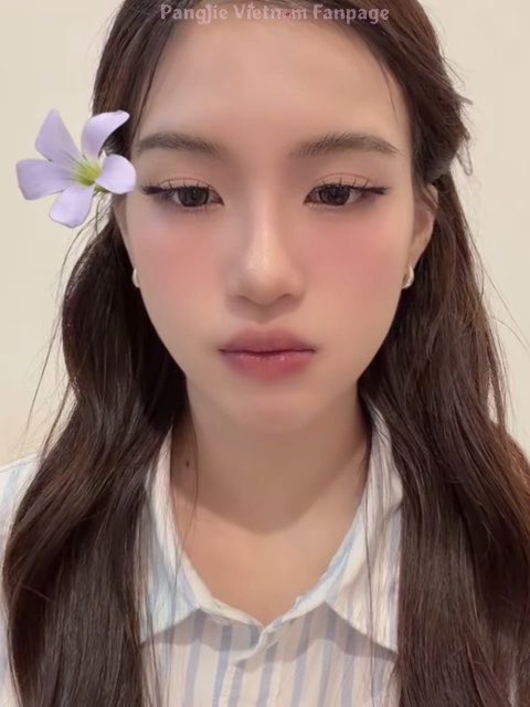 A close-up portrait of a young woman with long dark hair, pink blush makeup, and a purple flower tucked behind her ear, wearing a striped shirt, with watermark text 'Pangjie Vietnam Fanpage'.