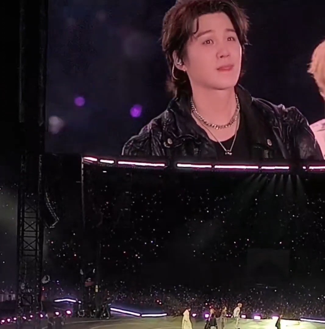 A young man wearing a black leather jacket and chain necklaces appears to be emotional, displayed on a large screen at a concert with a starry backdrop and a visible audience.