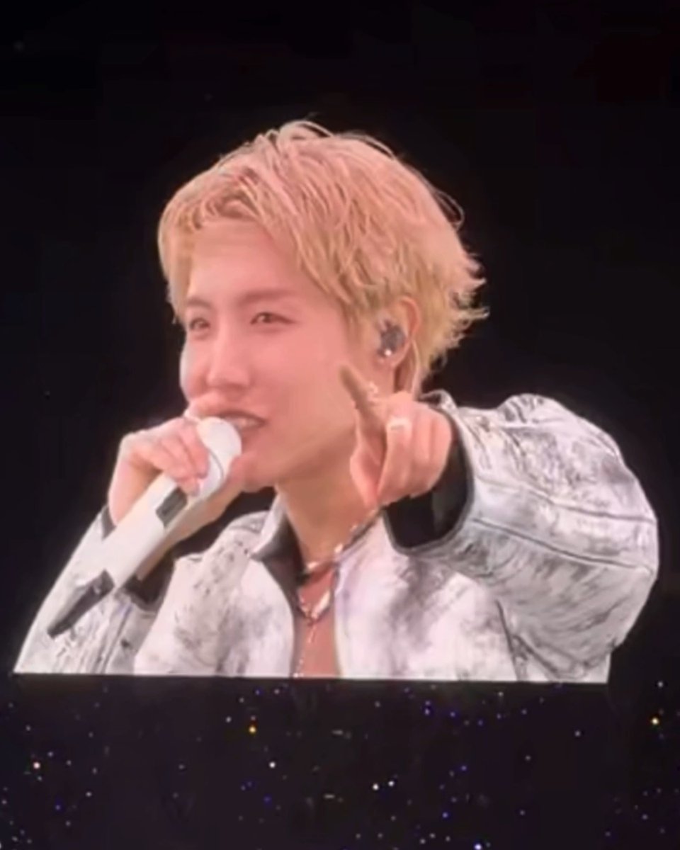 A blonde-haired K-pop performer on stage smiling while pointing toward the camera, holding a white microphone and wearing a denim jacket, with an in-ear monitor visible.