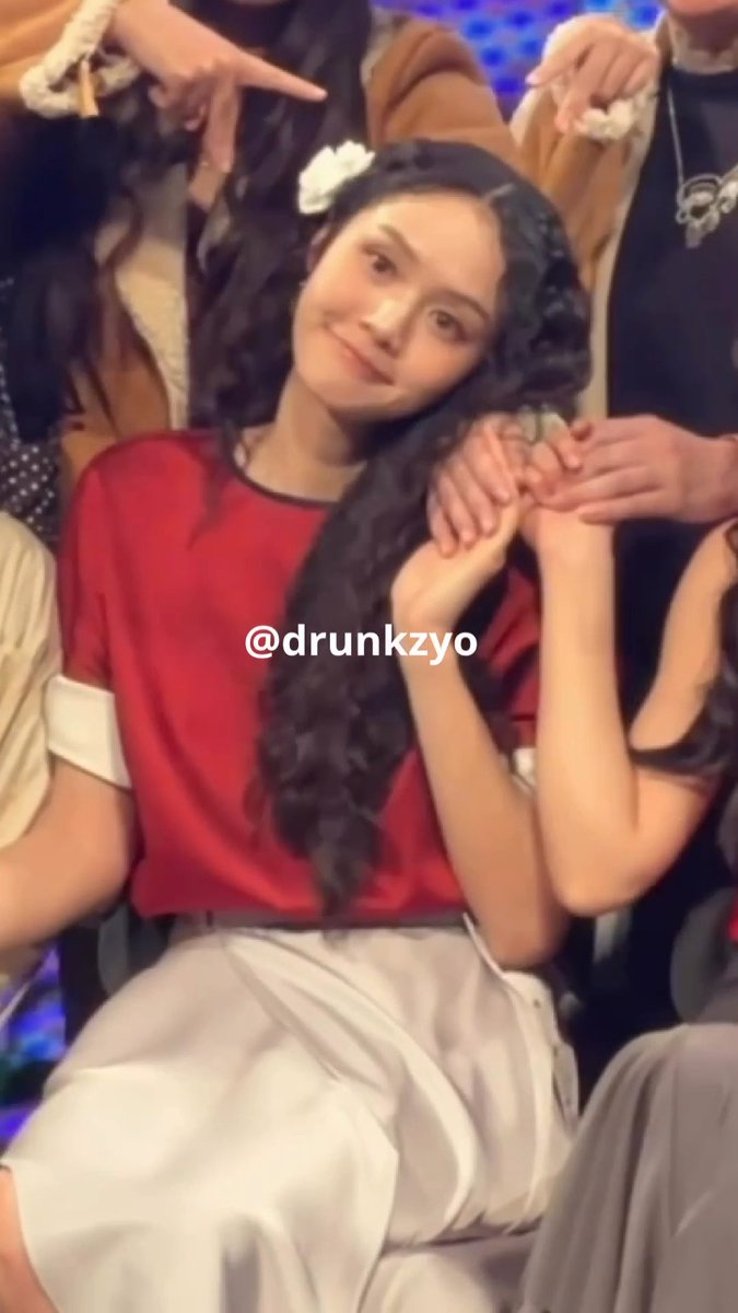 A photo of a smiling woman with long dark curly hair wearing a red top and white skirt, watermarked @drunkzyo.