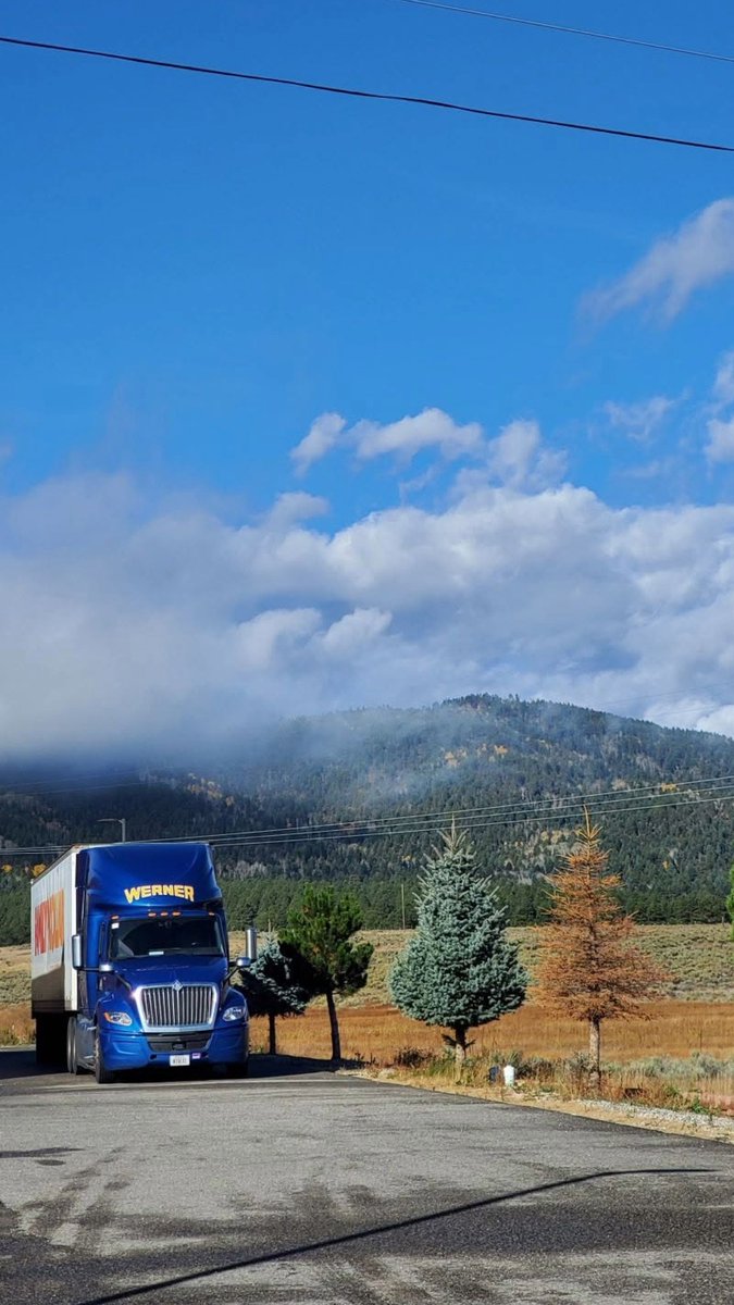 A blue semi-truck branded 'WERNER' is parked on a road against a scenic backdrop of mountains, clouds, and a vibrant blue sky.