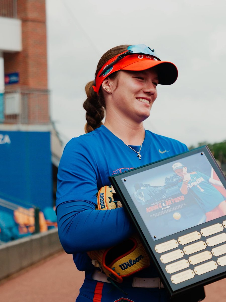 A smiling female athlete wearing a blue long-sleeve shirt and an orange visor holds a Wilson glove and a framed "Above & Beyond Award" plaque at what appears to be a softball stadium.