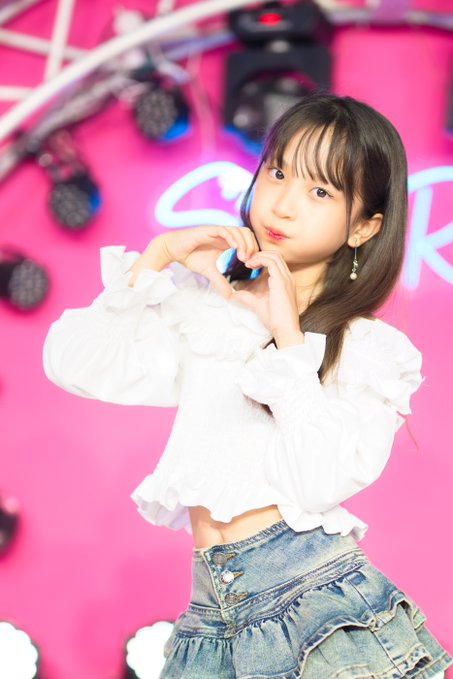 A young woman with long dark hair poses in a white ruffled cropped blouse and denim skirt, forming a heart shape with her hands against a bright pink backdrop with stage lighting.