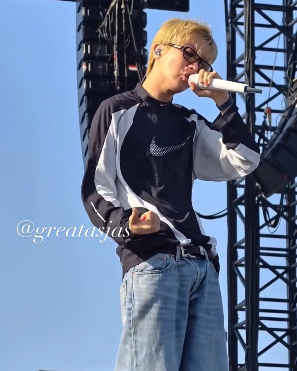 A blonde male performer wearing glasses and a black and white jacket singing into a microphone on stage, with a watermark reading '@greatasjas'.