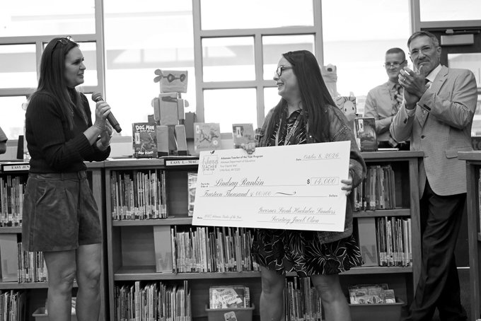 In a black-and-white photo, a woman holds a large presentation check for $13,000 made out to Lindsey Houkpin, while another woman speaks into a microphone and a man claps in front of a library bookshelf.