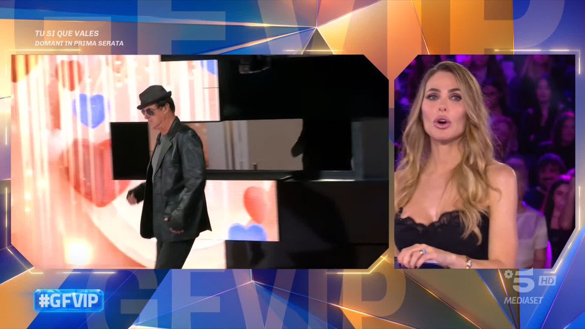 A split-screen broadcast from Italy's Mediaset channel 5 HD features "TU SI QUE VALES DOMANI IN PRIMA SERATA" text alongside "#GFVIP" branding, showing a man in a black hat and leather jacket on the left while a blonde female presenter in a black lace top reacts on the right, capturing the energetic reality TV moment being celebrated with "FLYING #GFVIP."
