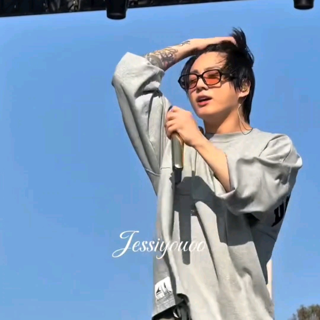 A young male performer with black hair and tinted sunglasses holds a microphone on stage against a clear blue sky, with a watermark reading 'Jessigouco'.
