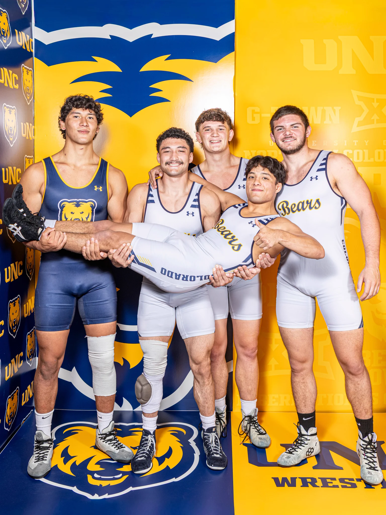 A group of five athletes in wrestling attire pose in front of a blue and yellow University of Northern Colorado (UNC) backdrop. Four are standing, smiling, while collectively holding up another teammate horizontally. The backdrop features UNC Bears logos.