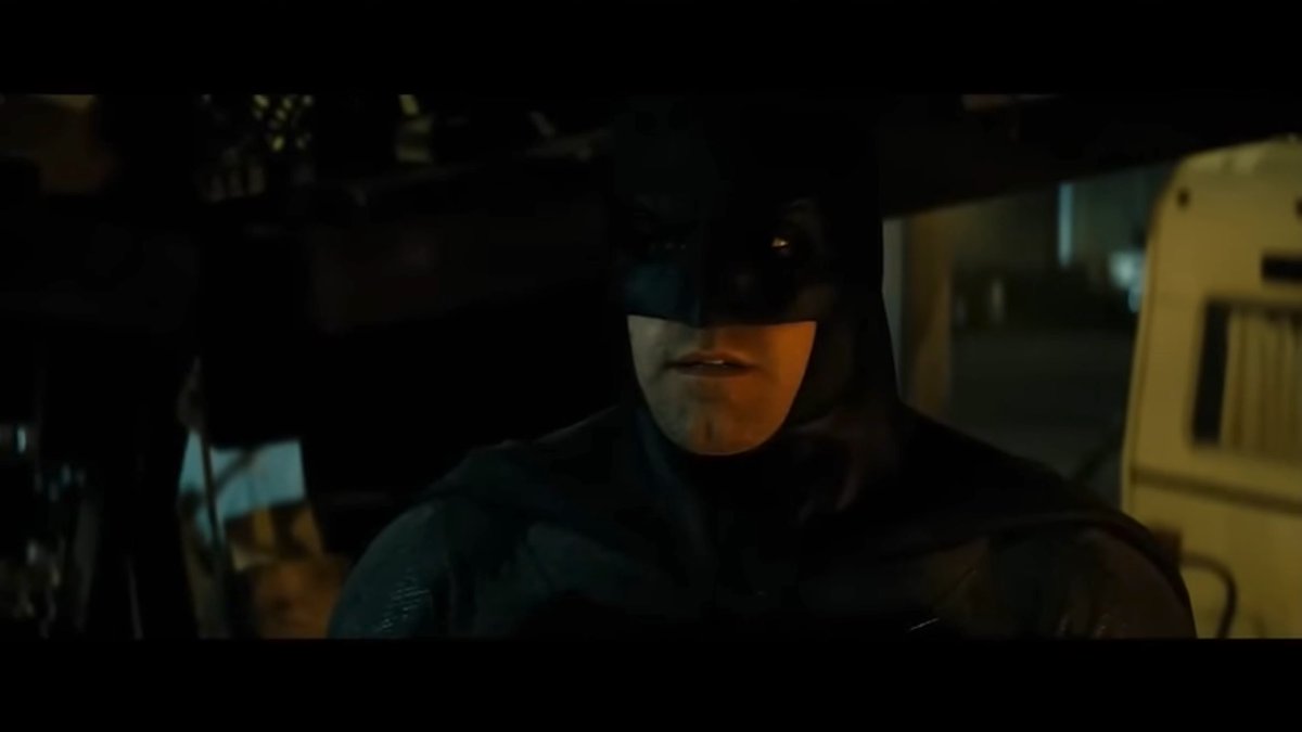 A cinematic shot of Batman in his cowl and suit, standing in a dimly lit, shadowy environment.