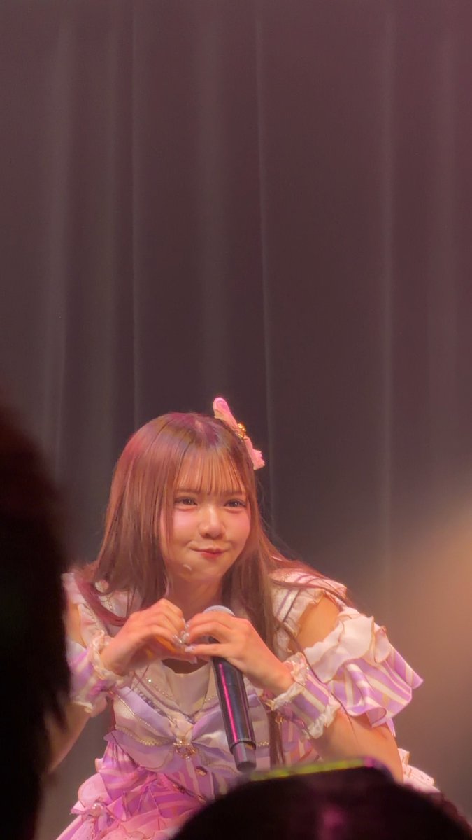 A young woman with long brown hair in a pink and white ruffled dress holding a microphone on stage, making a heart shape with her fingers.
